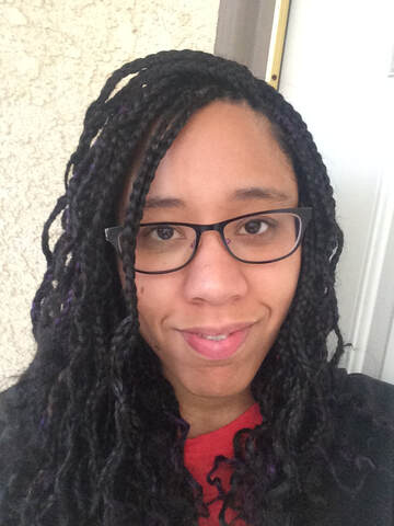 a black woman smiles at the camera. she has black and purple braids. Her hair is down and she wears glasses. 